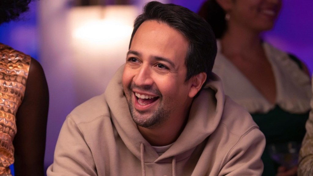 Lin-Manuel Miranda sits at a table and smiles in Percy Jackson.